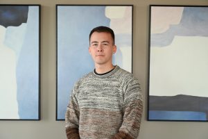 Alvin Osterndorff standing in front of three paintings wearing a sweater.