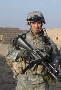 Brian Bieniek in a military uniform and protective gear standing outside while holding a rifle.