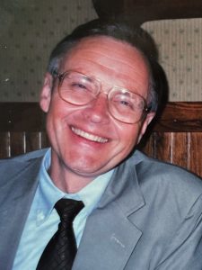 Dennis Carlson poses for a headshot in a gray suit with a black tie and glasses, smiling.