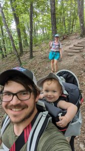 Joseph Miller carries a baby on his back while hiking through a forest, with another person in the background holding up a peace sign.