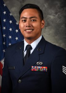 Lance Lazarte poses for a headshot in his Air Force uniform against a gray background with an American flag in the background.