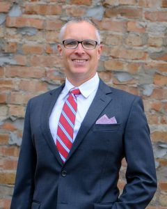 Matt Choquette poses for a headshot in a suit with a red and blue striped tie.