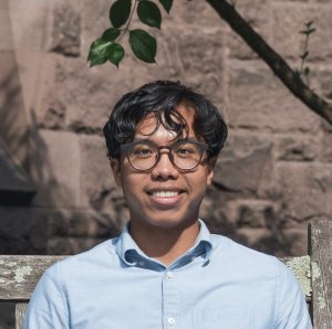 Miguel Pingol poses for a headshot wearing glasses and a blue collared shirt in front of a red stone wall.