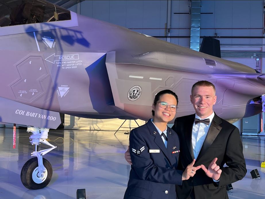 Thi Tran in her uniform and a man in a suit pose for a photo holding up a "W" with their hands together in front of a fighter jet.