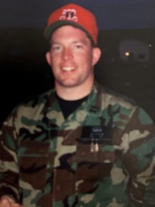 Todd Nelson smiling in a blurry photo taken at night. He is in his camoflauge uniform and a red baseball hat.
