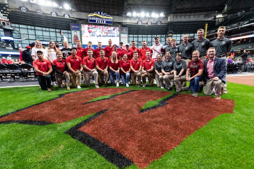 A group of UW-Madison military connected students students pose for a picture at the American Family Field in front of a large red "W" for Wisconsin Badgers.
