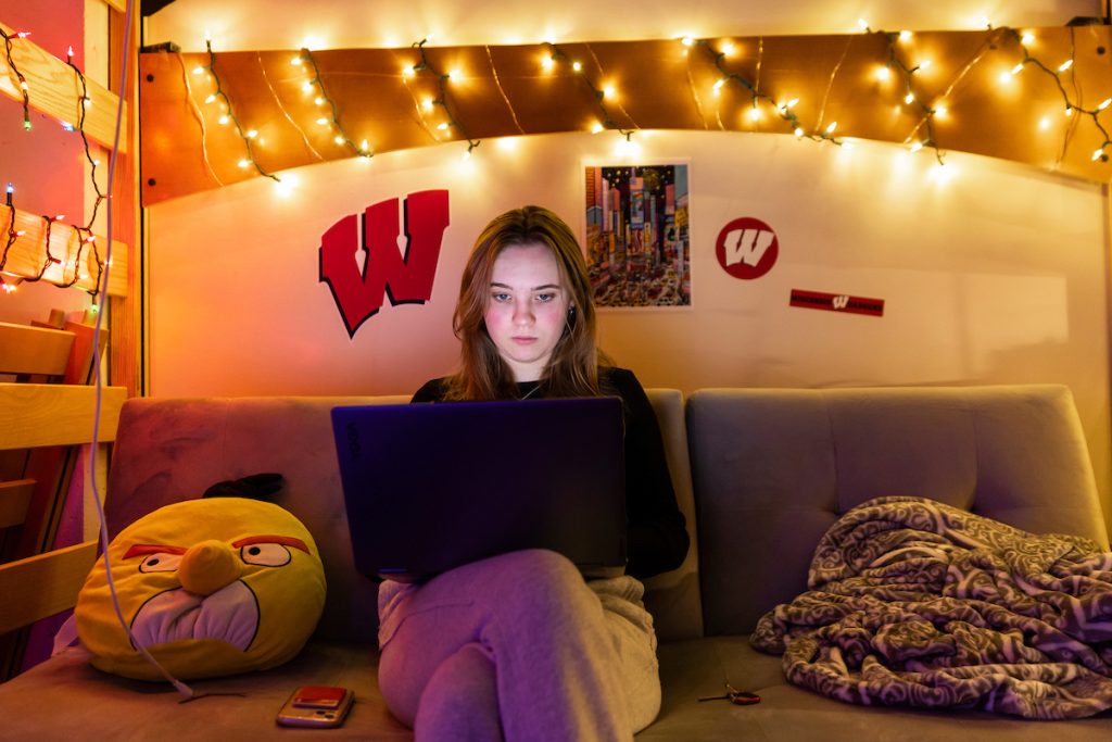 A woman in a black sweater and grey sweatpants sitting on a futon surrounded by Christmas lights looking at her laptop.