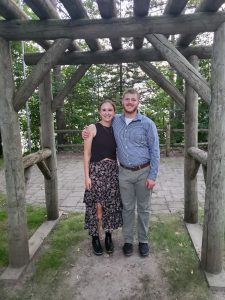 Nick Brennan and a woman wearing a black shirt and floral skirt posing for a picture under a wooden arch outside.