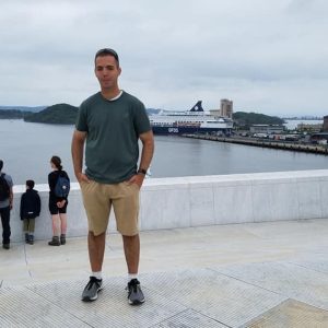 Zach Gerras standing by a river along a pier with a large boat docked in the background.