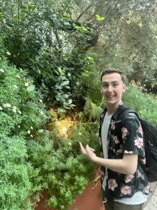 Jacob Warner wearing a tropical button-up t-shirt pointing at a bunch of green plants.