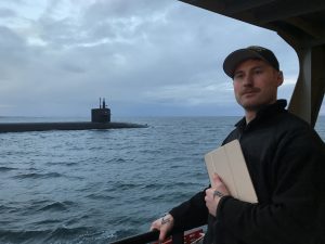 Andrew Ledoux holding an iPad with a body of water and submarine in the background.