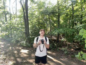 Owen Olichwier holding a camera on a hiking trail smiling for a photo.