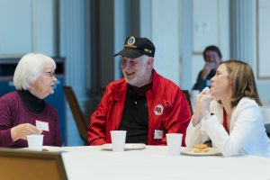 A man in a red jacket and black Vietnam veteran hat sitting at a table between two women. The woman on the right is wearing a white shirt and the woman on the left is wearing a purple sweater. They are smiling and laughing as the woman in purple talks.