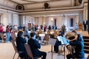 Five brass musicians sitting in chairs by a stage playing music. The rest of the room is filled with tables and people standing with their hands over their chests as they face the front of the room.