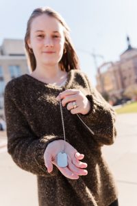 A woman with blond hair and wearing a brown sweater stands outside. She has her hand outstretched holding a military dog tag in one hand and the chain in her other hand.