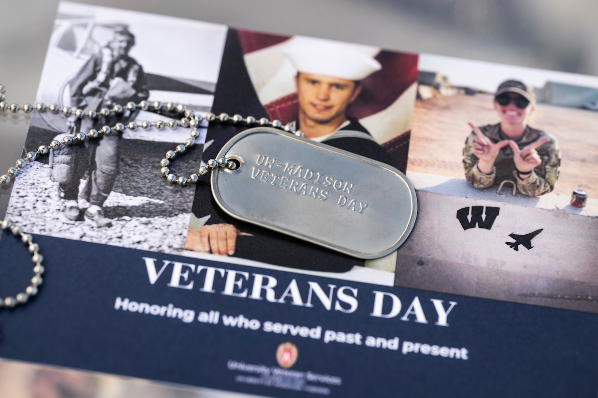 A military dog tag and chain set on top of a postcard with three pictures of soldiers in uniform. The postcard says, "Veterans Day: Honoring all who served past and present"