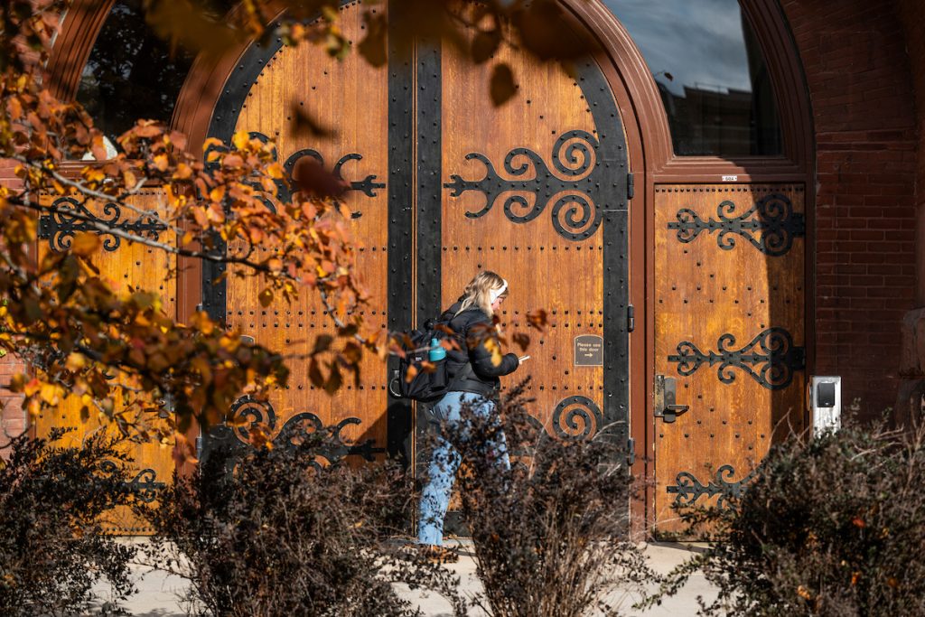 A pedestrian walks by as tree leaves frame a view of the main doors to the Red Gym (Armory and Gymnasium) at the University of Wisconsin–Madison during a late autumn day.